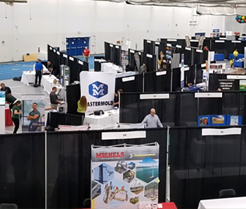 Indoor job or trade fair with multiple booths separated by black curtains. Several people are walking and talking at different booths displaying banners and promotional materials. The event appears organized and busy.