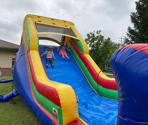 Two young girls are sliding down a large, colorful inflatable slide outdoors on a grassy area, with buildings and trees in the background under a cloudy sky.