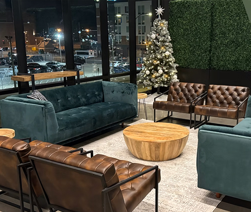 A modern lounge area with green velvet sofa, brown leather armchairs, round wooden coffee tables, a light-colored rug, and a decorated Christmas tree near large windows overlooking a city at night.