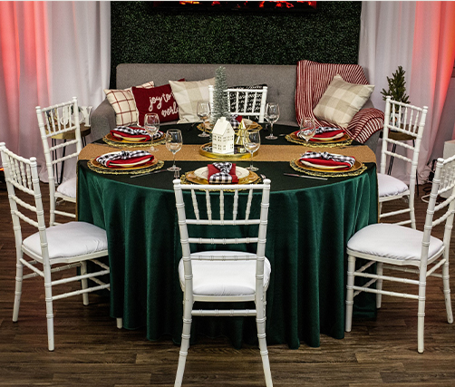 A round dining table with a dark green tablecloth is set for six with festive plates, napkins, and glasses. White chairs surround the table, and a gray sofa with red and plaid pillows is in the background.