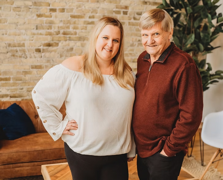 A woman in a white off-the-shoulder top stands next to an older man in a maroon sweater. Both are smiling, posing indoors with a brick wall, sofa, and plant in the background.