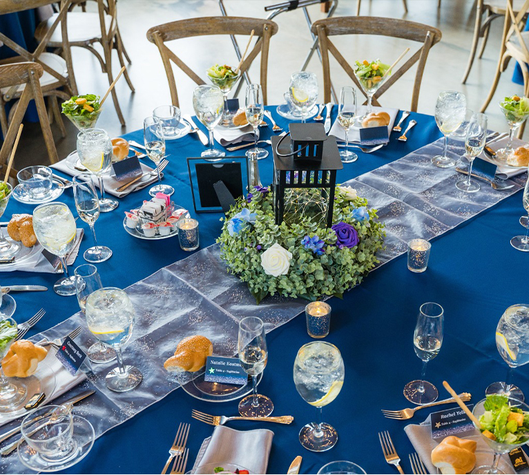 A round table set for a formal event with blue tablecloth, wooden chairs, floral centerpiece with white and purple roses, glassware, salad plates, bread rolls, and place cards at each setting.