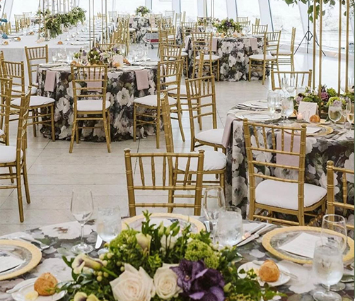 Elegant wedding reception with round tables covered in floral-patterned tablecloths, gold Chiavari chairs, floral centerpieces, and place settings. The room is bright with natural light and decorated for a formal event.