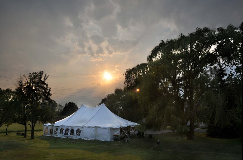A large white event tent stands on a grassy field surrounded by trees at sunset, with dramatic clouds and soft sunlight filtering through the sky.