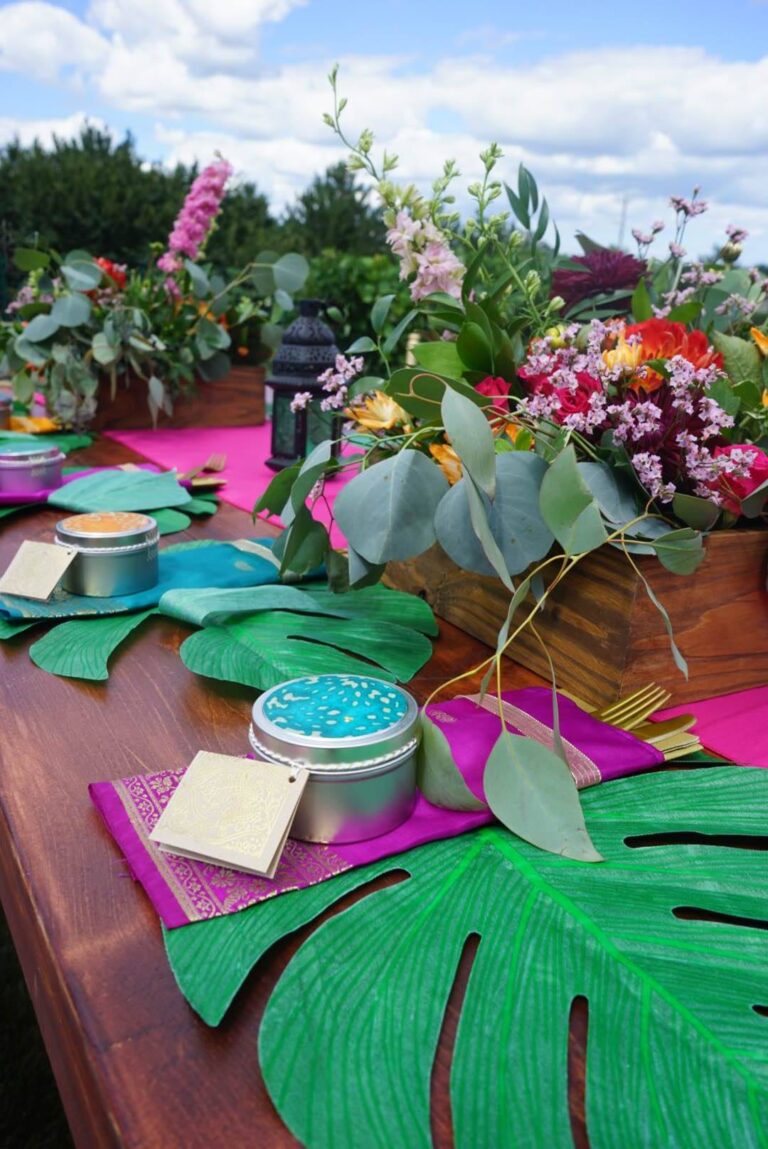 A colorful outdoor table setting with tropical leaf placemats, tins with teal lids, bright pink table runners, and vibrant flower arrangements in wooden boxes under a blue sky with clouds.