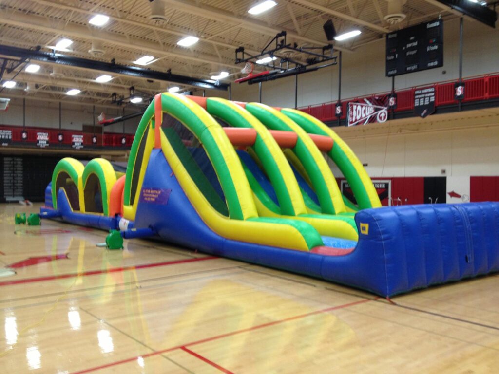 A large, colorful inflatable obstacle course is set up inside a gymnasium, featuring arches, slides, and climbing sections on a polished wooden floor with red and black gym decor in the background.
