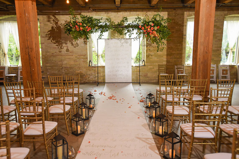 A wedding ceremony setup with gold chairs in rows, a white aisle runner with script, black lanterns with candles lining the aisle, and a floral arrangement of greenery and orange flowers hanging above the altar area.