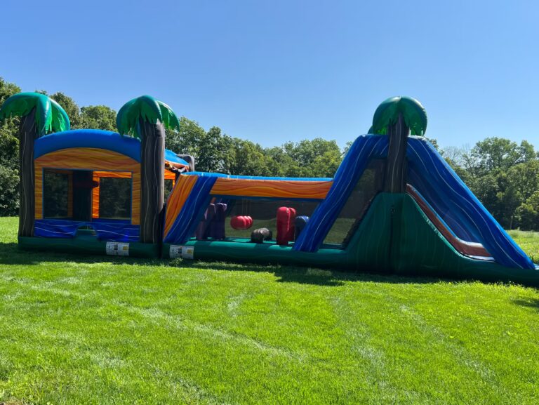 An inflatable obstacle course with palm tree decorations sits on bright green grass under a clear blue sky, with trees in the background.