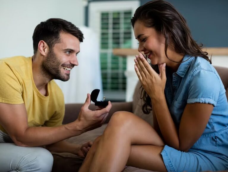 A man kneels and presents an open ring box to a smiling, surprised woman sitting on a couch, suggesting a marriage proposal in a cozy indoor setting.