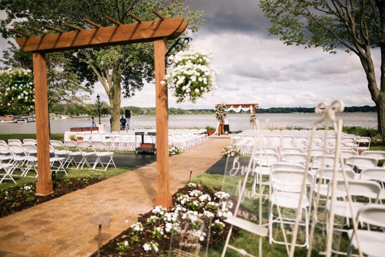 An outdoor wedding ceremony setup by a lake, featuring rows of white chairs, a wooden pergola with hanging flowers, and a decorative archway at the end of a brick aisle under cloudy skies.