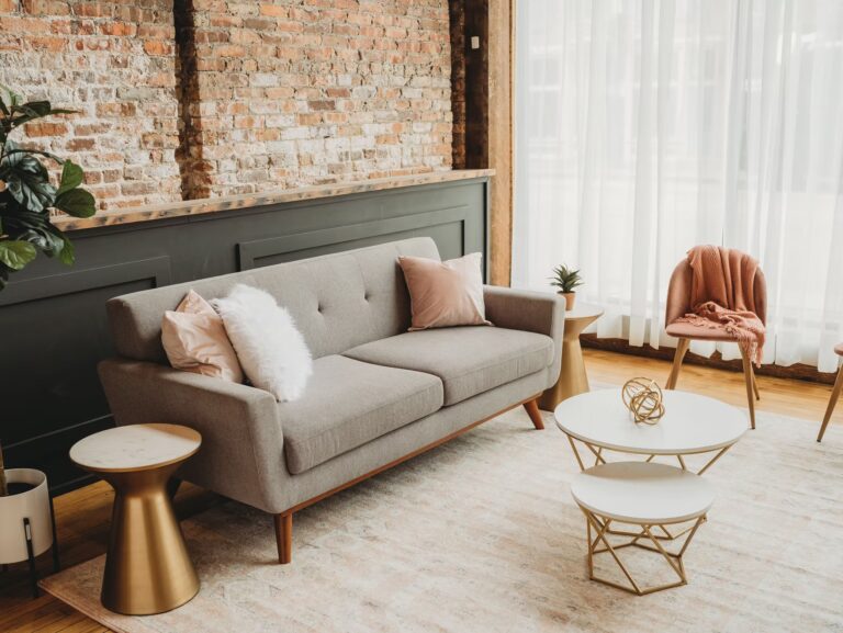 A modern living room with a gray sofa, pink and white pillows, round coffee tables, a pink chair with a throw blanket, and a brick accent wall. Sunlight streams in through sheer white curtains.