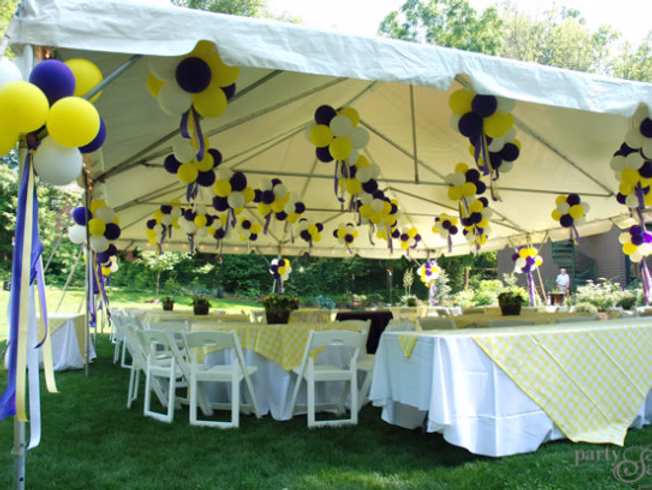 A white outdoor event tent with white tables and chairs, decorated with yellow, purple, and white balloons and ribbons. The tables are covered with yellow plaid tablecloths, set on green grass with trees in the background.