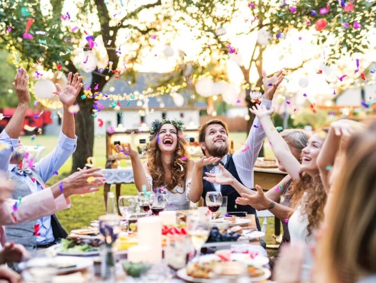 A group of people sitting around a decorated outdoor table, joyfully tossing colorful confetti into the air, celebrating together in a lively garden setting.