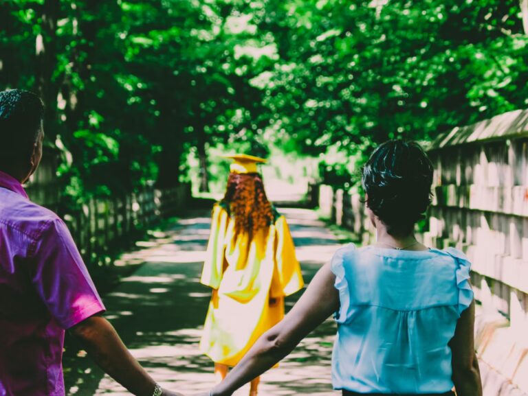 Two adults hold hands in the foreground while watching a young woman in a yellow graduation gown and cap walk down a tree-lined path away from them. Sunlight filters through the leaves above.
