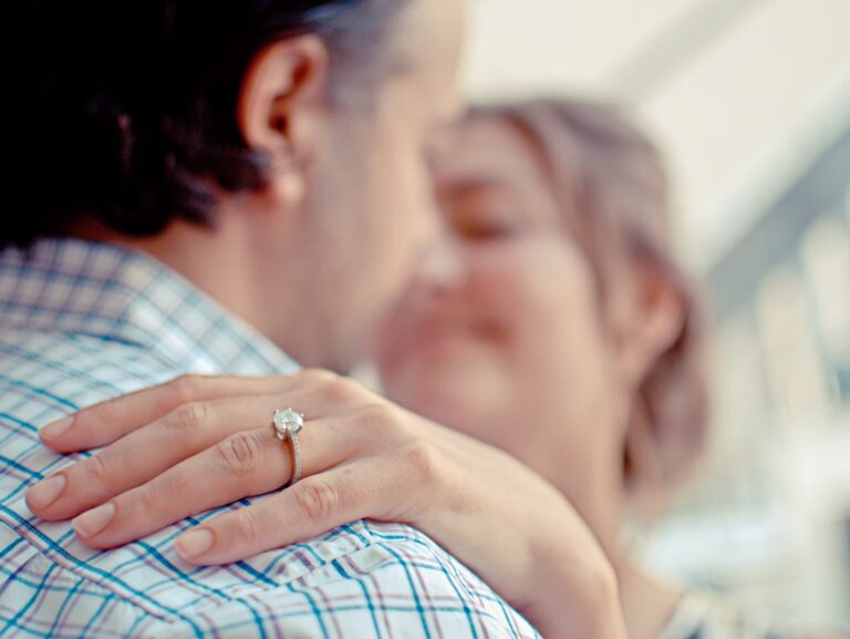 A close-up of a couple embracing, with focus on a womans hand resting on the mans shoulder, showing an engagement ring with a large diamond. Their faces are softly blurred in the background.