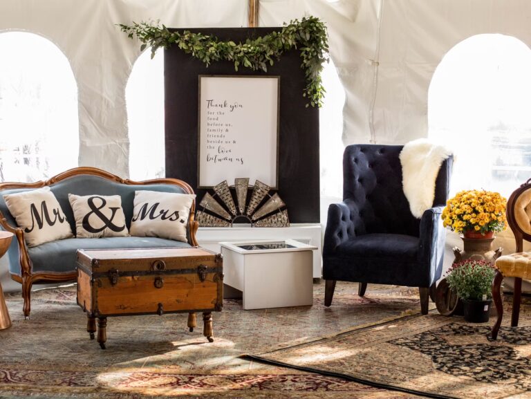 Elegant living room setup with a vintage sofa featuring Mr. & Mrs. pillows, a navy blue armchair with a fur throw, a wooden chest as a coffee table, flowers, and decorative framed sign with greenery above.