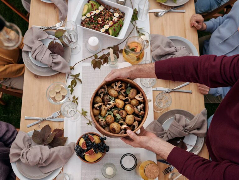 A person holds a bowl of roasted potatoes and mushrooms over a wooden table set for a meal, with various dishes, drinks, and decorative napkins arranged around.