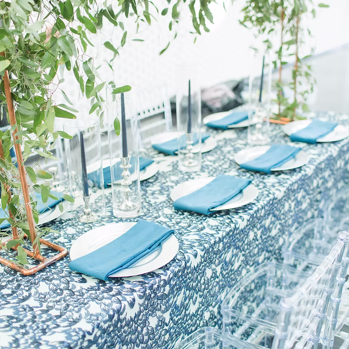 A table set for a formal event features blue-patterned tablecloth, white plates with neatly folded blue napkins, clear glasses, tall candlesticks, and overhead greenery creating a fresh, elegant atmosphere.