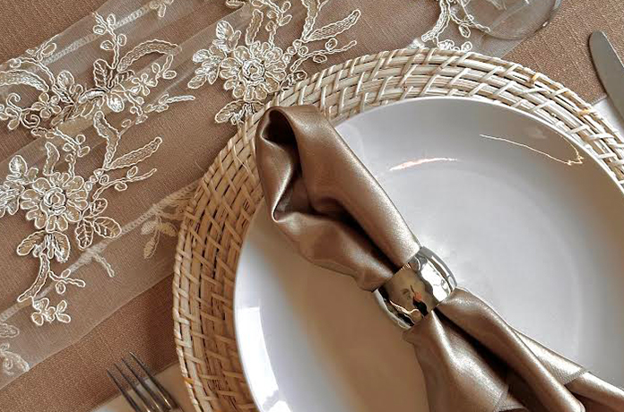 A close-up of an elegant table setting with a white plate on a woven placemat, a metallic napkin ring holding a folded satin napkin, cutlery, a wine glass, and a lace table runner with floral patterns.