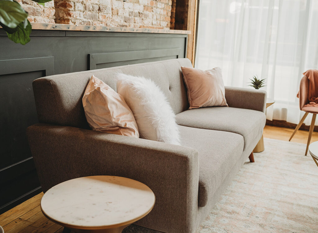 A modern gray sofa with blush pink and white fluffy pillows sits in a cozy living room with a round marble side table, exposed brick wall, and large sheer curtains letting in natural light.