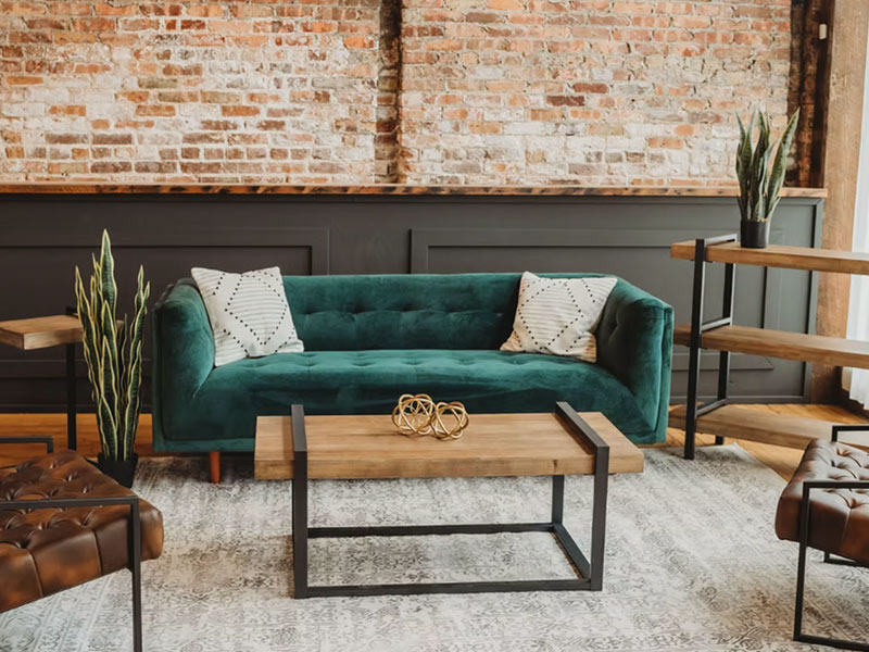A modern living room with a green velvet sofa, two geometric pillows, a wooden coffee table with metal legs, plants, and exposed brick wall in the background. Shelves and leather seating are also visible.