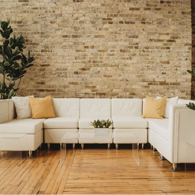 A modern living room with a white sectional sofa, yellow throw pillows, a small glass coffee table with a plant, wood flooring, and a light brown exposed brick wall in the background.