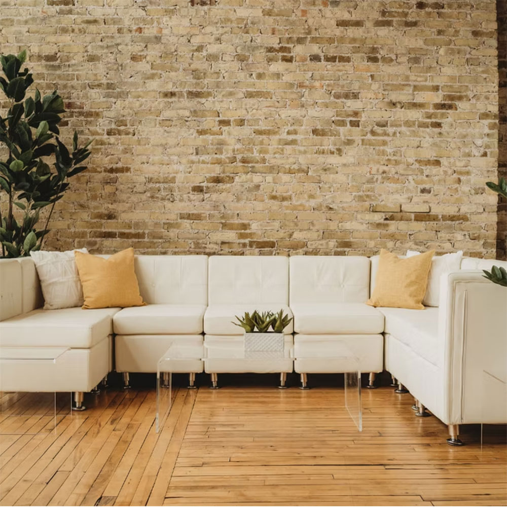 A modern living room with a white sectional sofa, yellow throw pillows, a small glass coffee table with a plant, wood flooring, and a light brown exposed brick wall in the background.