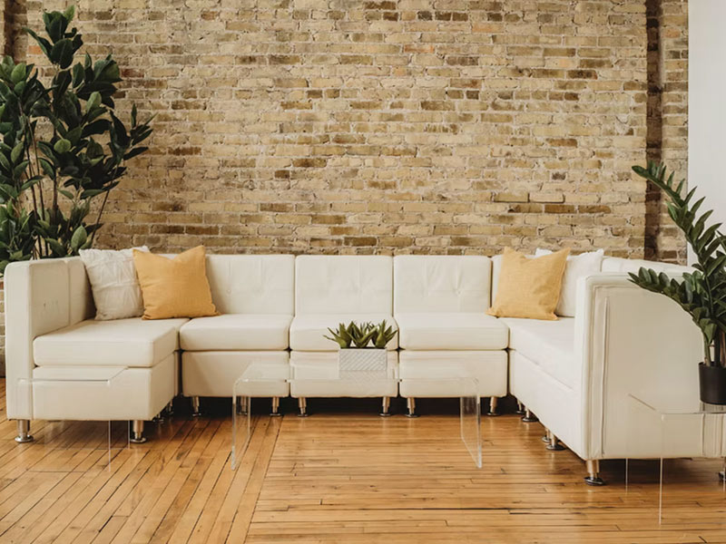 A modern living room with a white sectional sofa, yellow throw pillows, clear acrylic tables, green potted plants, a wooden floor, and an exposed brick wall.