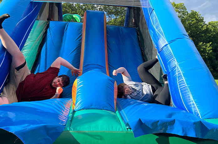 Two people are sliding down a large blue inflatable slide outdoors, landing at odd angles and laughing, with trees visible in the background on a sunny day.