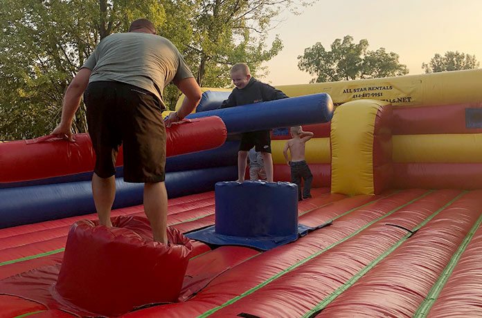 Two children and an adult play on an inflatable jousting arena outdoors. The adult and one child hold large padded sticks, while another child stands nearby. Trees and sunlight are visible in the background.