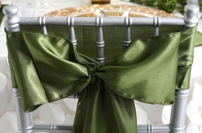 A silver chair with a shiny green satin ribbon tied in a bow around the backrest, set in front of a table with a decorative plate and white tablecloth.