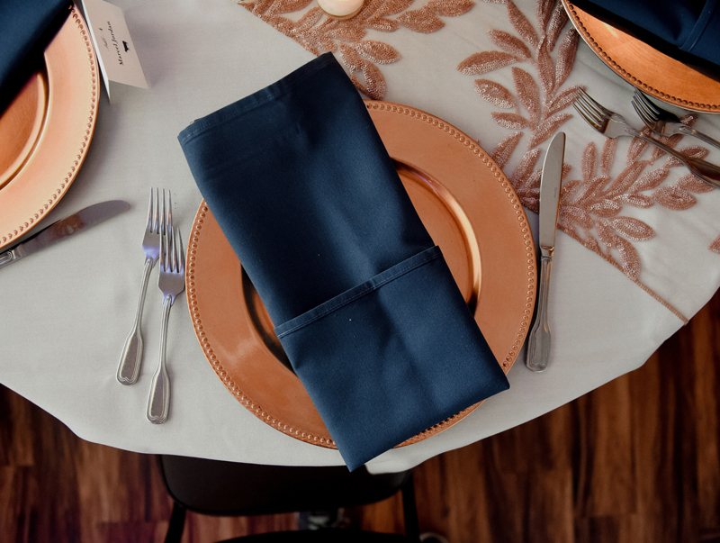 A table setting with a gold-rimmed plate, navy blue napkin, silver cutlery, and a tablecloth with a shimmery leaf design. The image is taken from above, showing part of a wooden floor.