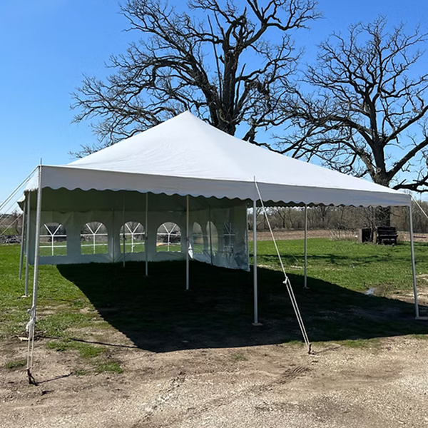 A large white outdoor event tent with open sides stands on grass under a clear blue sky, with bare trees and a field visible in the background.