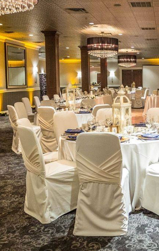 A banquet hall set for an event, featuring round tables covered with white tablecloths and chairs draped in white covers, elegant chandeliers, candles, and soft ambient lighting.