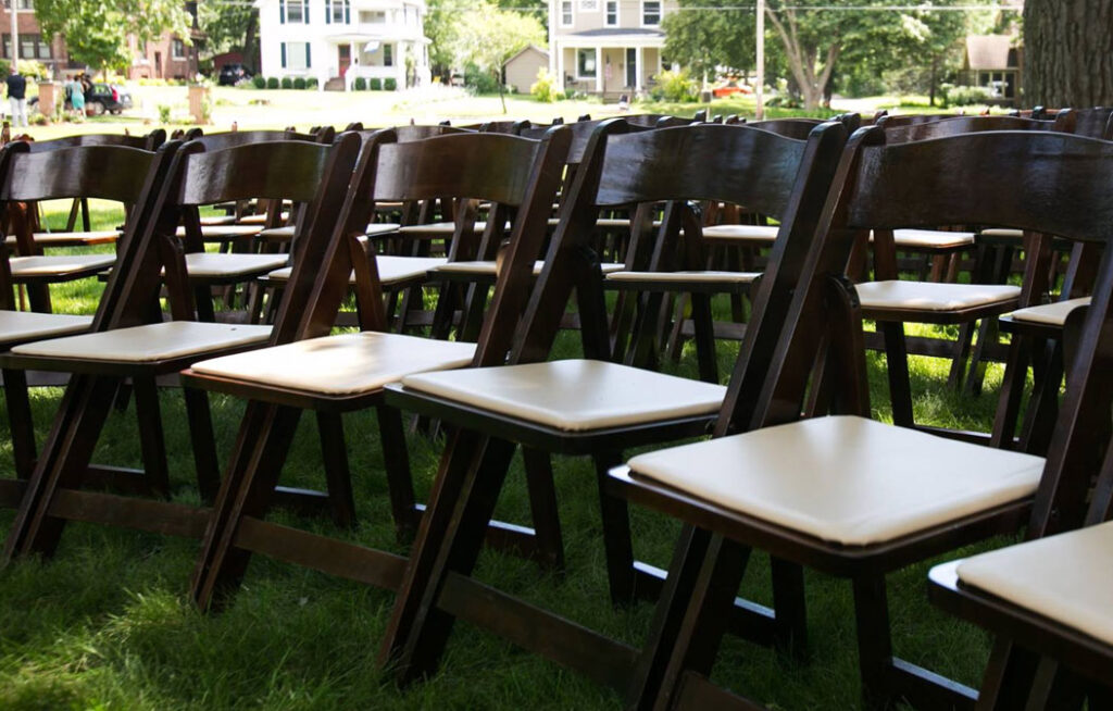 Rows of empty wooden chairs with white cushions set up outdoors on green grass, likely for an event or ceremony, with houses and trees visible in the background.