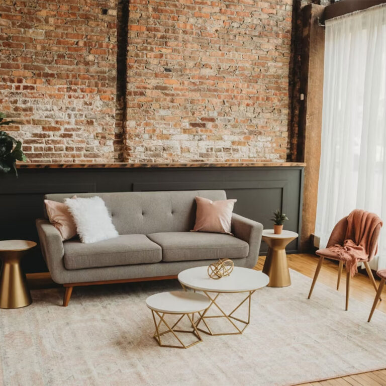 A modern living room with a grey sofa, pink and white pillows, a pink throw on a chair, gold side tables, geometric coffee tables, a plant, brick wall, and large sheer curtains letting in natural light.