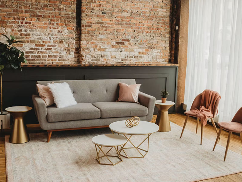 A modern living room with a gray sofa, pink and white cushions, gold accent tables, two pink chairs, a potted plant, and large windows with sheer white curtains. The backdrop features a brick wall.