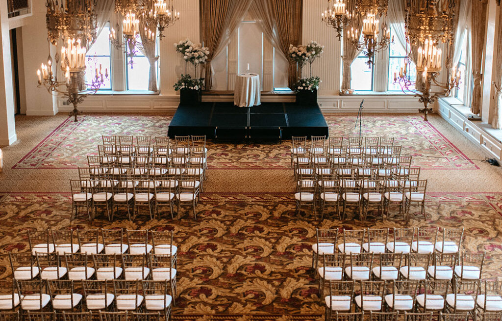 An elegant indoor wedding venue with rows of chairs facing a small stage, two large chandeliers, floral arrangements, and tall windows with curtains letting in natural light.