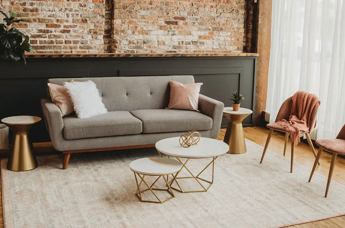A modern living room with a gray sofa, pink and white pillows, two gold side tables, nested white coffee tables with gold bases, a pink chair, and a brick accent wall. Light streams in through sheer curtains.