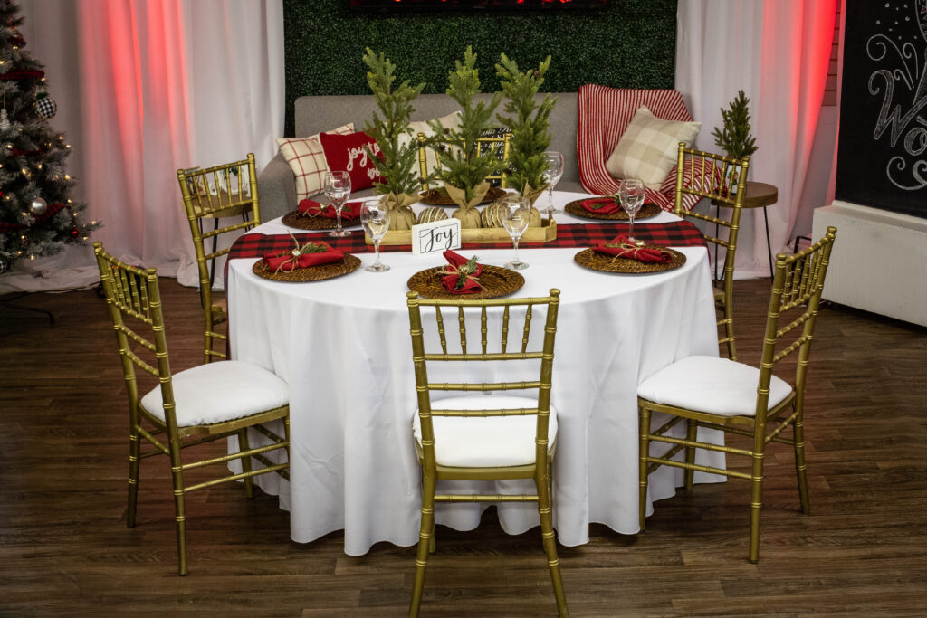 A round table set for six with gold chairs, festive red and green decor, mini Christmas trees, and place settings. The background has a Christmas tree, couch with pillows, and holiday decorations.