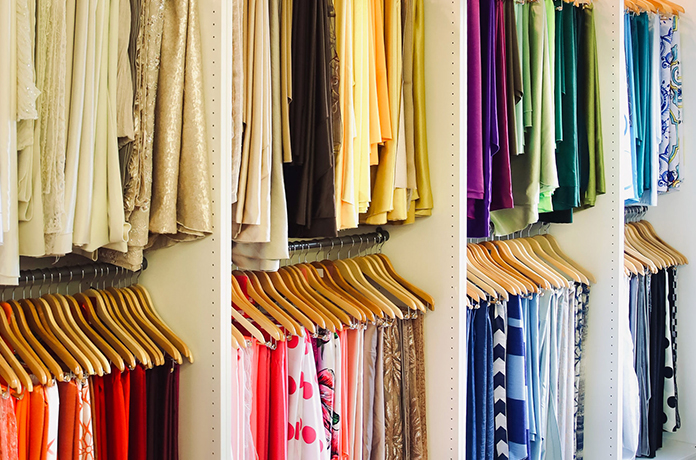 Clothing is neatly arranged on wooden hangers and shelves, organized by color from warm reds and yellows to cool blues and greens, creating a visually appealing rainbow effect in a wardrobe or closet.