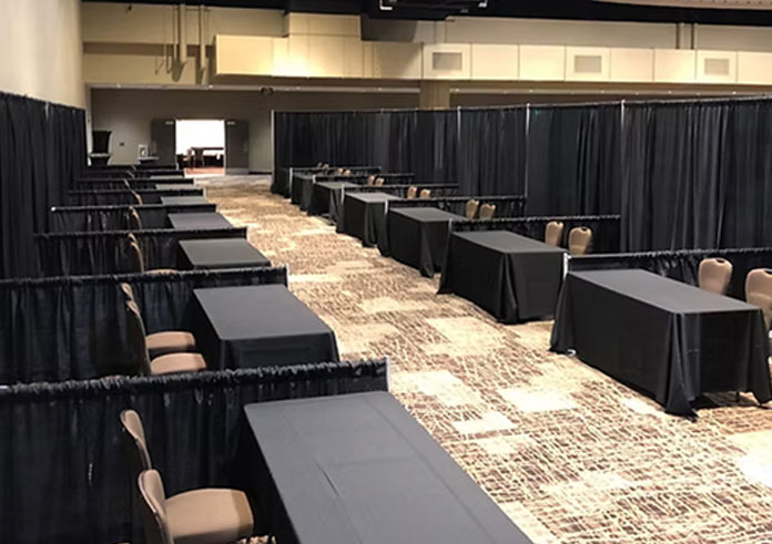 A large room with rows of tables covered in black tablecloths, separated by black curtains, and surrounded by empty chairs, resembling a setup for an event or convention.