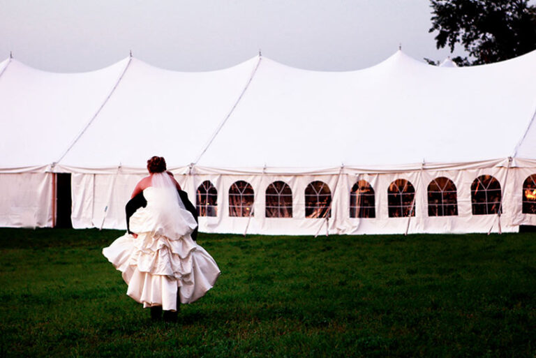 A bride in a white wedding gown and veil walks across a grassy field toward a large white event tent with arched windows.