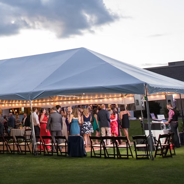 A group of people dressed formally gather under a large white event tent with string lights at an outdoor evening party. Empty black chairs are arranged around the tent on the grass.