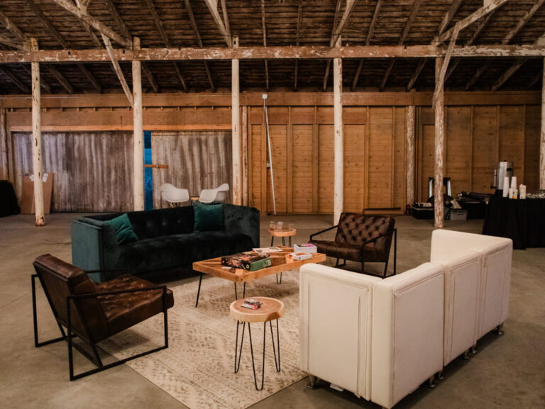 A cozy lounge area in a rustic barn features modern sofas, a coffee table with board games, side tables, and a rug. Exposed wooden beams and unfinished walls add to the barn’s industrial charm.