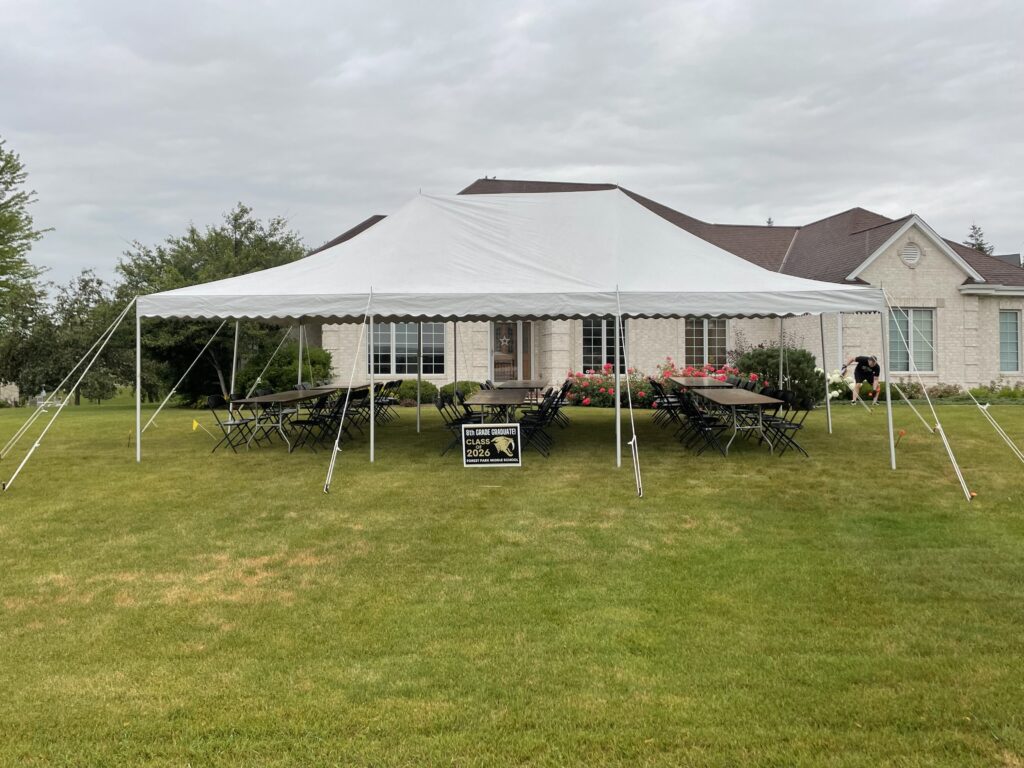 A large white tent with folding tables and chairs set up underneath on a green lawn in front of a light-colored house, ready for an outdoor event. A sign and a few people are visible near the tent.