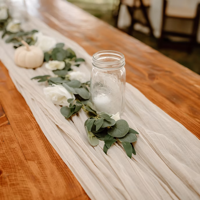 A wooden table is decorated with a light, flowing table runner, green eucalyptus leaves, white flowers, a small white pumpkin, and a clear mason jar holding a white candle.
