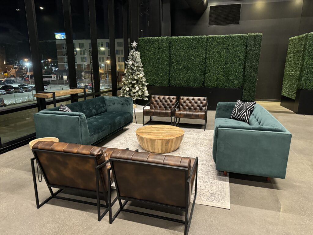 Modern lounge area with green velvet sofas, brown leather chairs, a round wooden coffee table, a light rug, and a decorated Christmas tree, set against tall windows and artificial green hedges.
