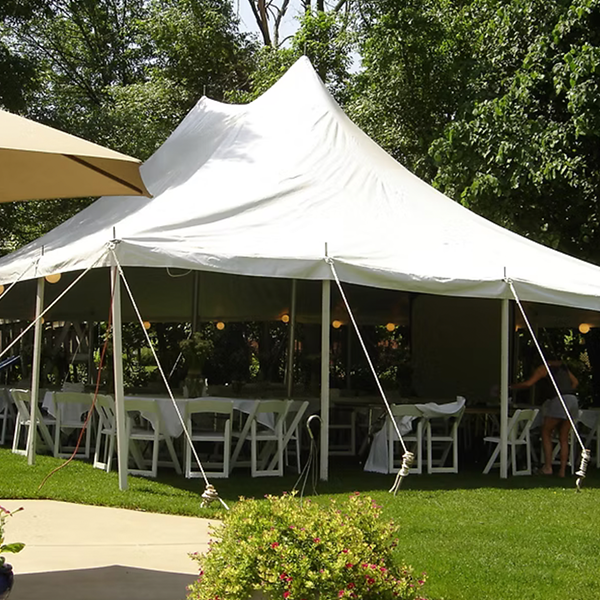 A large white event tent with a peaked roof is set up outdoors on a green lawn, sheltering rows of white chairs and tables draped in tablecloths; trees and plants surround the space.