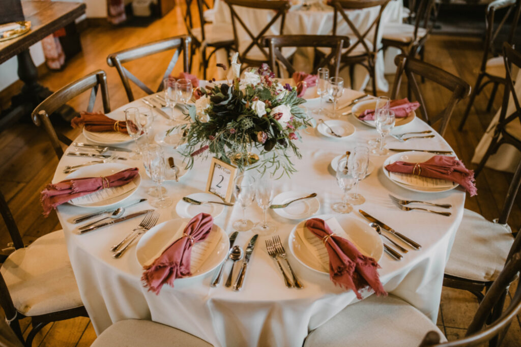 A round table set for eight with white tablecloth, pink napkins, plates, cutlery, wine glasses, and a floral centerpiece. A small sign with the number 2 is placed next to the arrangement.